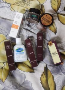 A collection of free sample products received from Smytten, displayed on a floral bedsheet. The items include: Deconstruct Vitamin C & Ferulic Acid Serum, mCaffeine Body Scrub, Pilgrim 24K Gold Facial Mask, Cetaphil Gentle Skin Cleanser, four Blush Pour Femme perfume vials (8ml each), and a Red Vine Face Serum.